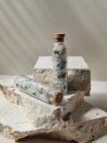 One vial of Rainbow Moonstone chips standing on a stone, a second lying on its side in front